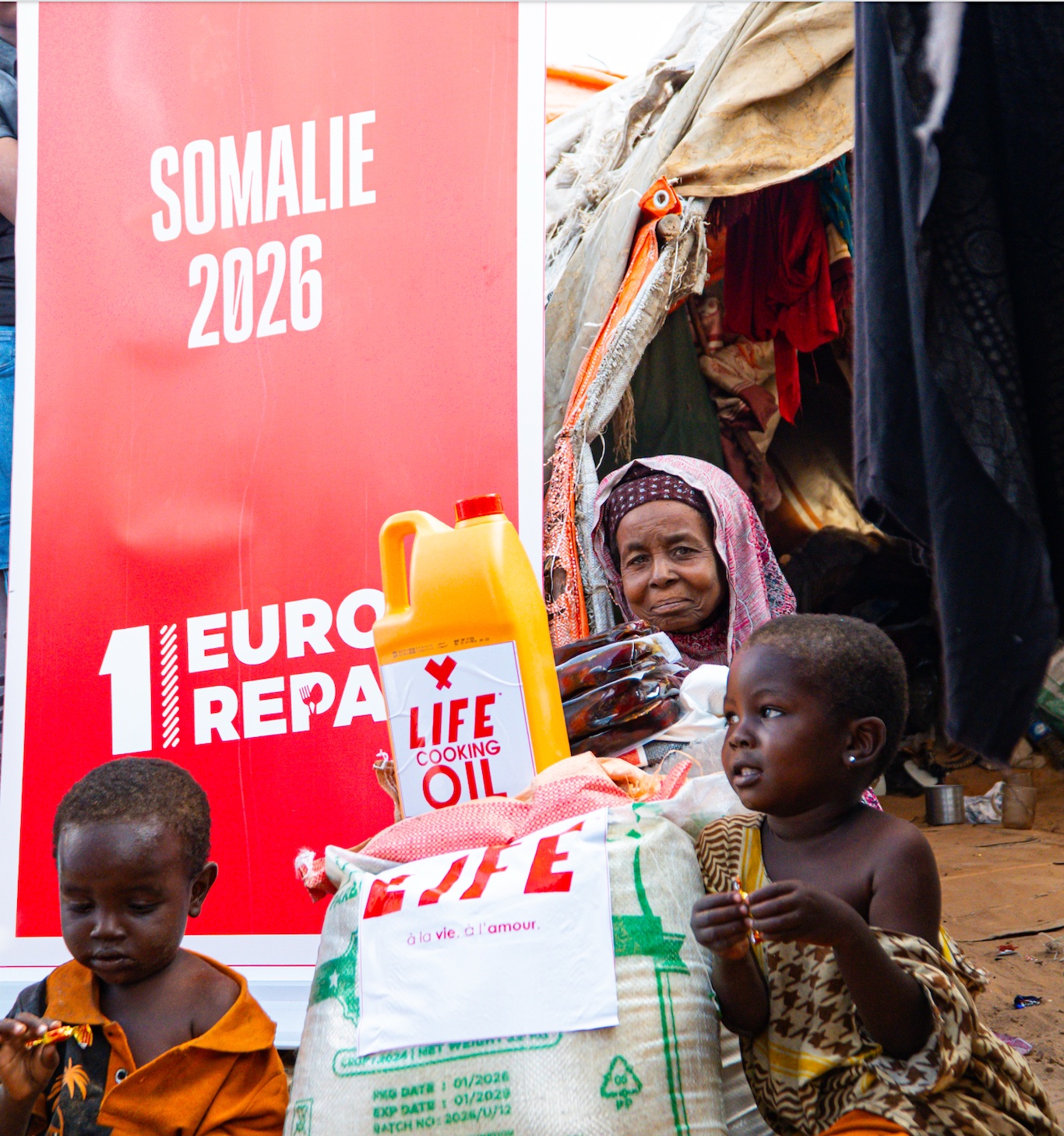 Deux enfants devant un sac de vivres et un bidon d'huile LIFE, une femme derrière eux, près d'une affiche Somalie