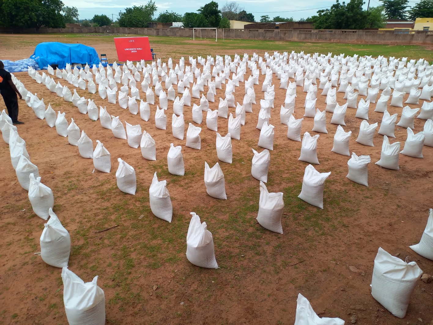 Sacs blancs alignés en rangées sur un terrain de terre, devant une bâche bleue et une affiche LIFE Urgence Mali.