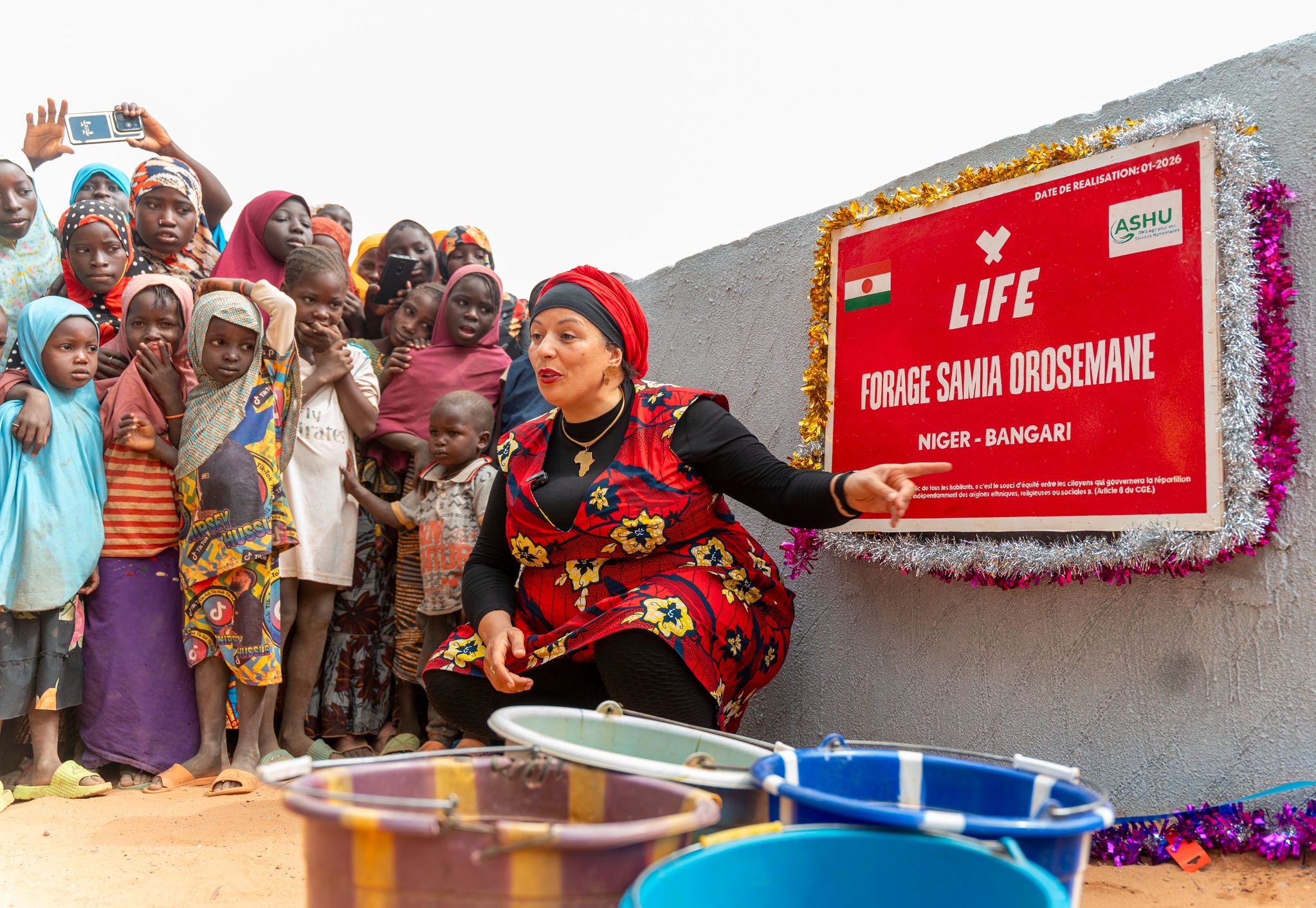 Femme accroupie devant un panneau LIFE « Forage Samia Orosemane, Niger - Bangari », entourée d'enfants et de seaux.