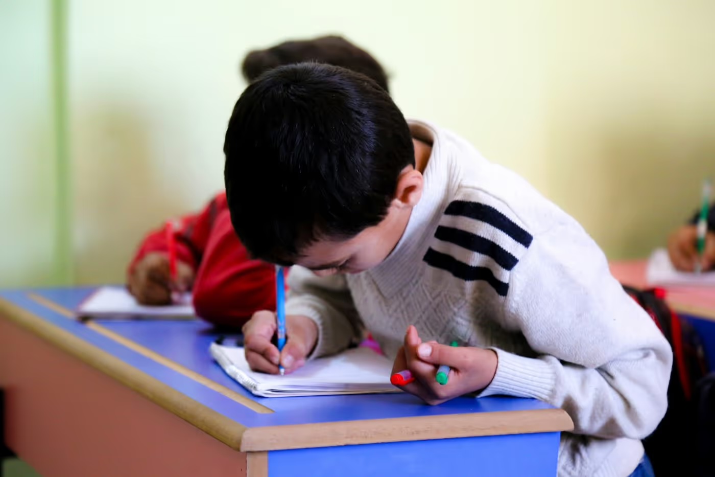 Un écolier penché sur son cahier écrit au stylo dans une salle de classe soutenue par LIFE.